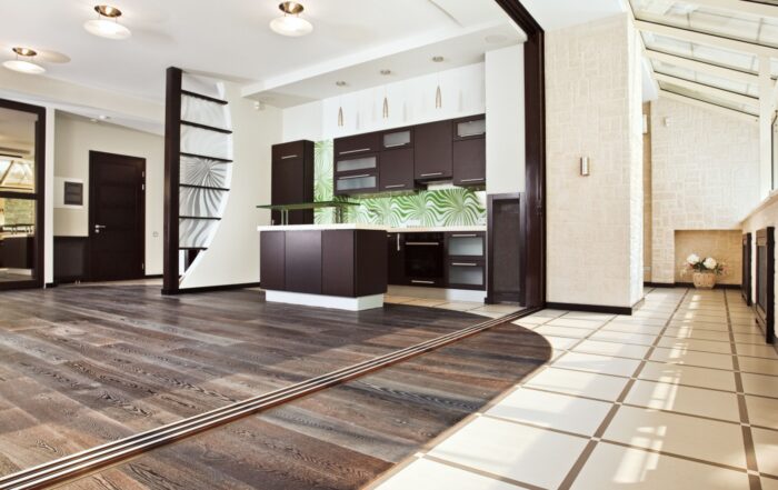 Modern open-concept kitchen and living area featuring a mix of dark hardwood flooring and light tile flooring. The hardwood floor adds warmth, while the contrasting tile creates a visually distinct area, enhancing the space's design and functionality. A glass backsplash with green patterns and ample natural light complement the elegant and stylish decor.