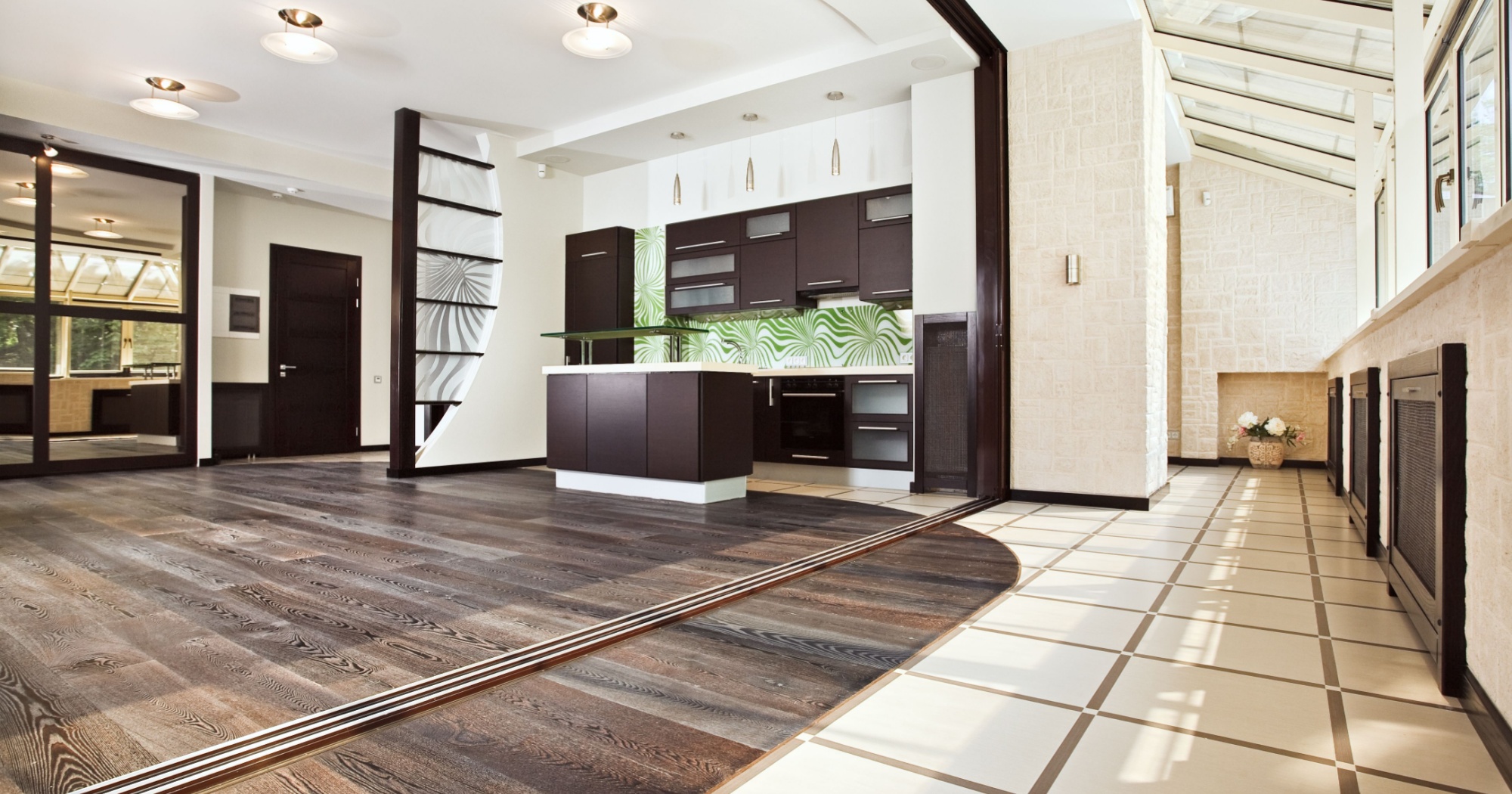 Modern open-concept kitchen and living area featuring a mix of dark hardwood flooring and light tile flooring. The hardwood floor adds warmth, while the contrasting tile creates a visually distinct area, enhancing the space's design and functionality. A glass backsplash with green patterns and ample natural light complement the elegant and stylish decor.