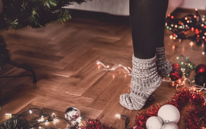 Cozy socks on a person standing near a Christmas tree with holiday decorations, lights, and tinsel on a polished wooden floor.