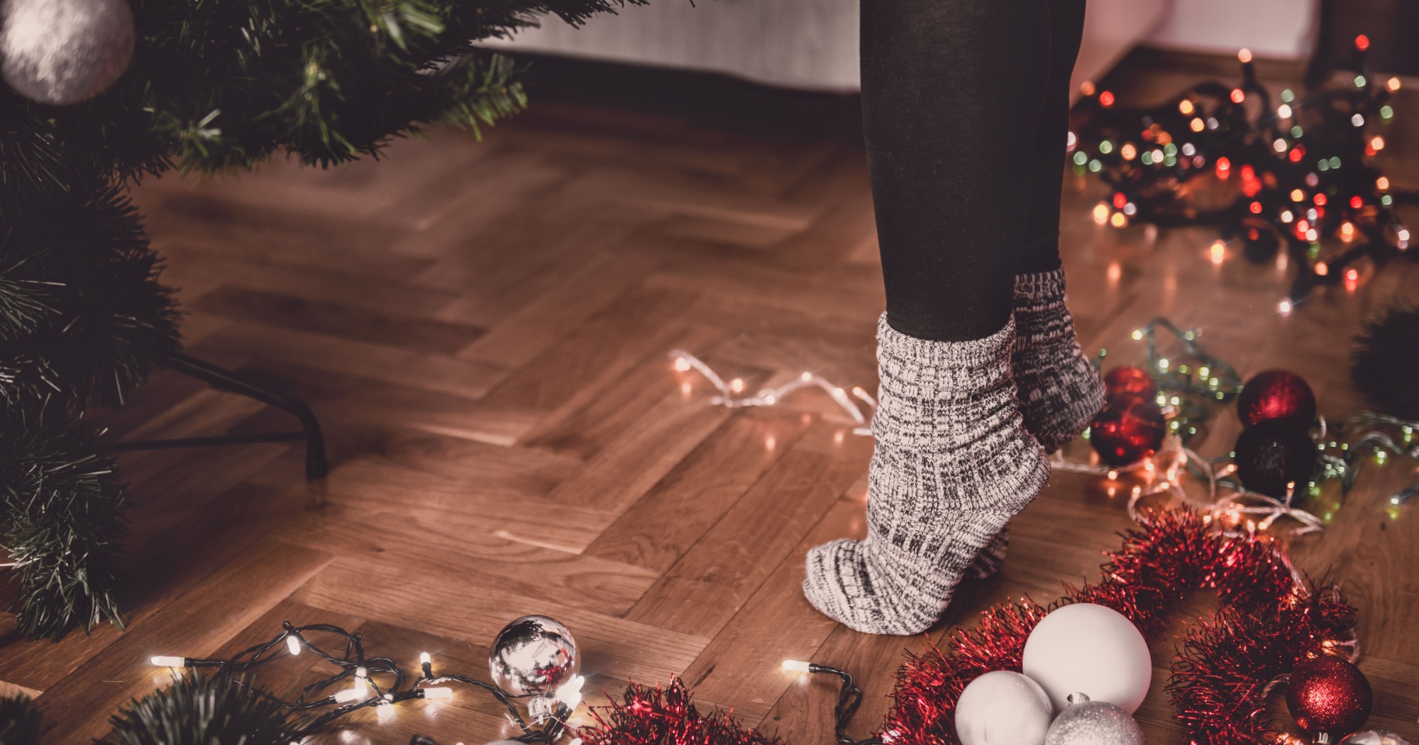 Cozy socks on a person standing near a Christmas tree with holiday decorations, lights, and tinsel on a polished wooden floor.